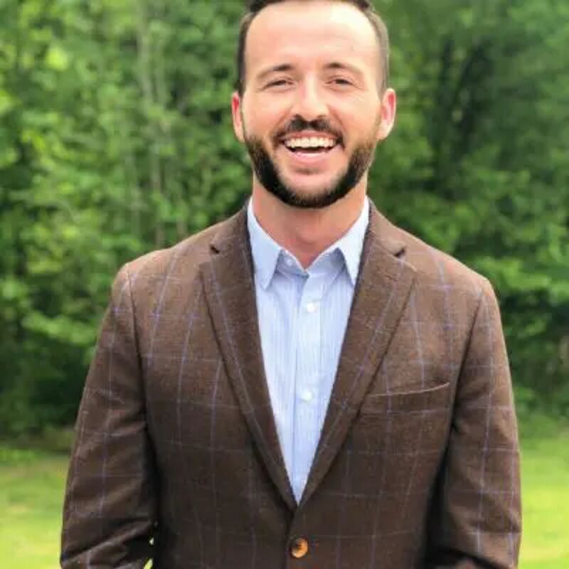 The image shows a man with a beard and short brown hair, wearing a brown blazer over a light blue shirt. He is smiling and looking directly at the camera. The background is a blurry green, suggesting an outdoor setting with trees or foliage. The man appears to be well-groomed and dressed in business casual attire.
